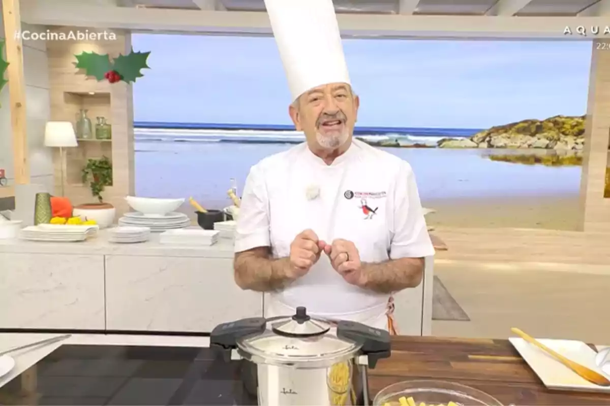 Karlos Arguiñano amb una gorra alta està en una cuina moderna amb una olla a pressió sobre la taula i un fons de platja a la pantalla. Captura de 'Cuina Oberta'