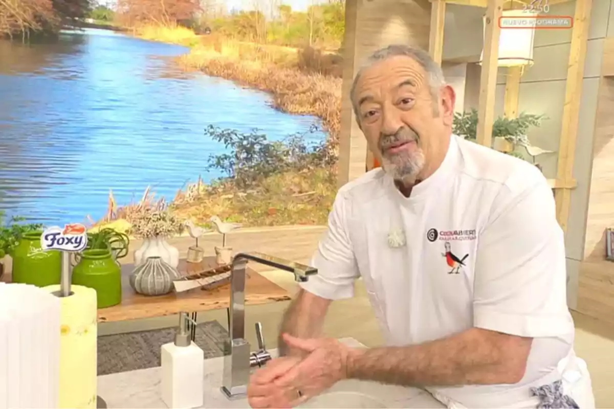 Karlos Arguiñano amb uniforme de xef s'està rentant les mans en una cuina amb un fons que mostra un paisatge de riu i vegetació. Captura de 'Cuina Oberta'.