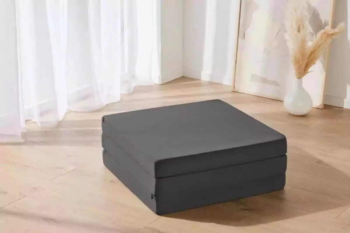 Un coixí quadrat gris fosc al terra d'una habitació amb terra de fusta clara i cortines blanques. Un coixí quadrat gris fosc al terra d'una habitació amb terra de fusta clara i cortines blanques.