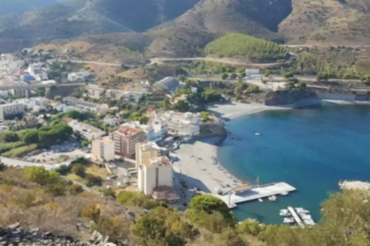 Vista aèria d´una ciutat costanera amb edificis, platja i un port petit, envoltada de muntanyes i vegetació.