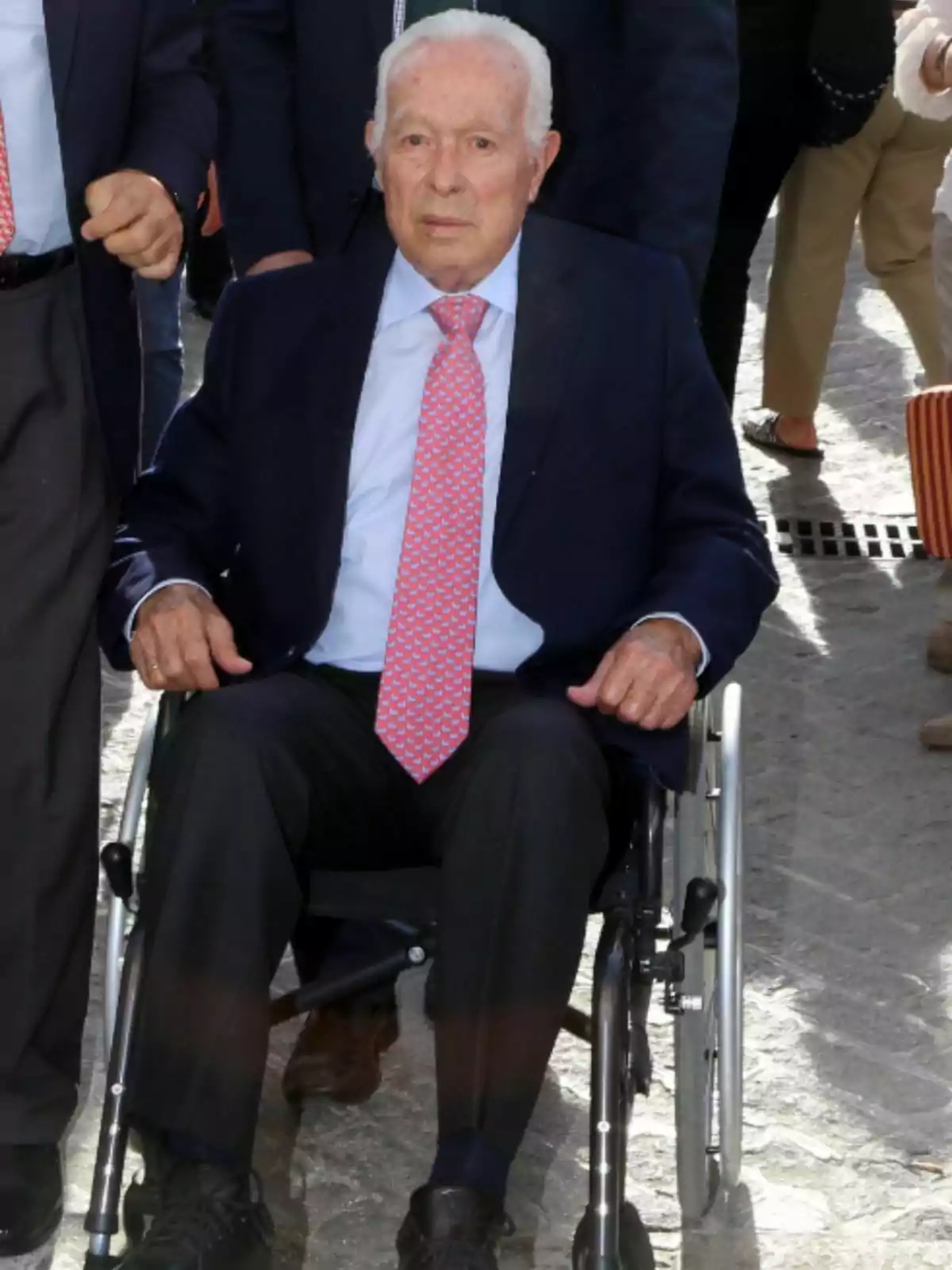 Curro Romero en cadira de rodes, vestit amb vestit fosc i corbata rosa, envoltat de persones. Curro Romero en cadira de rodes, vestit amb vestit fosc i corbata rosa, envoltat de persones.