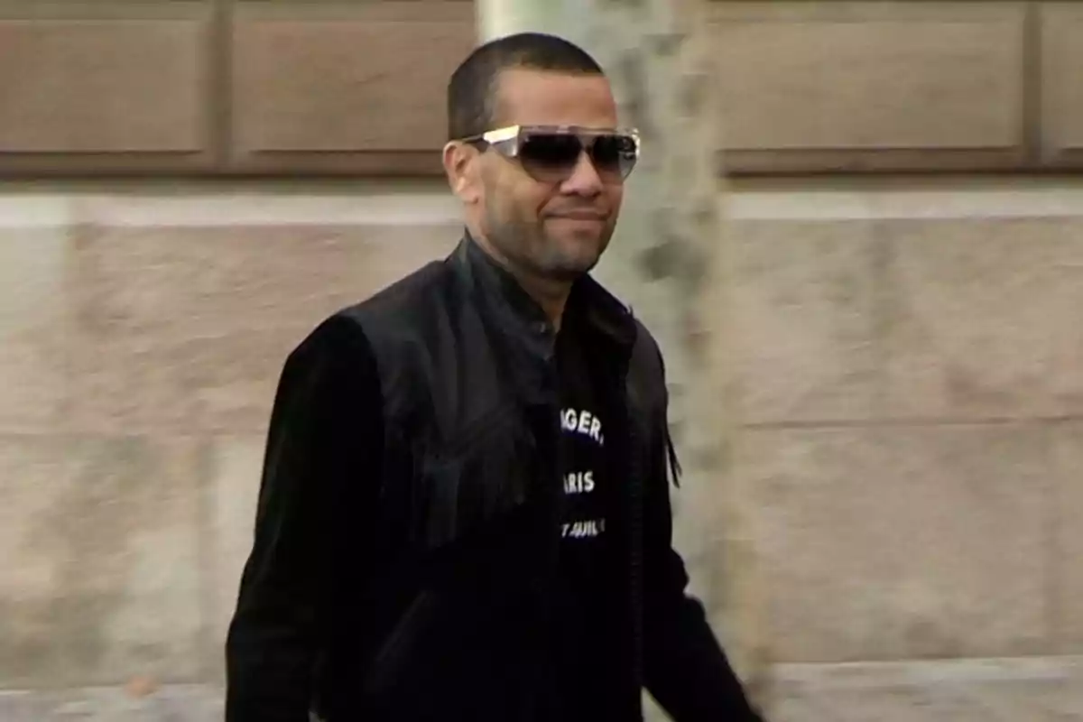 Dani Alves amb ulleres de sol i jaqueta negra camina a l'aire lliure davant d'una paret de pedra. Dani Alves amb ulleres de sol i jaqueta negra camina a l'aire lliure davant d'una paret de pedra.