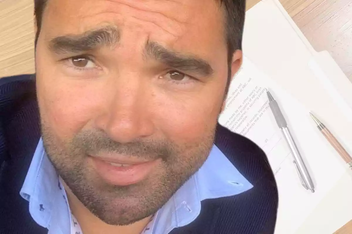 A man with a beard and expressive eyebrows looks at the camera, with documents and pens on a wooden table in the background. A man with a beard and expressive eyebrows looks at the camera, with documents and pens on a wooden table in the background.