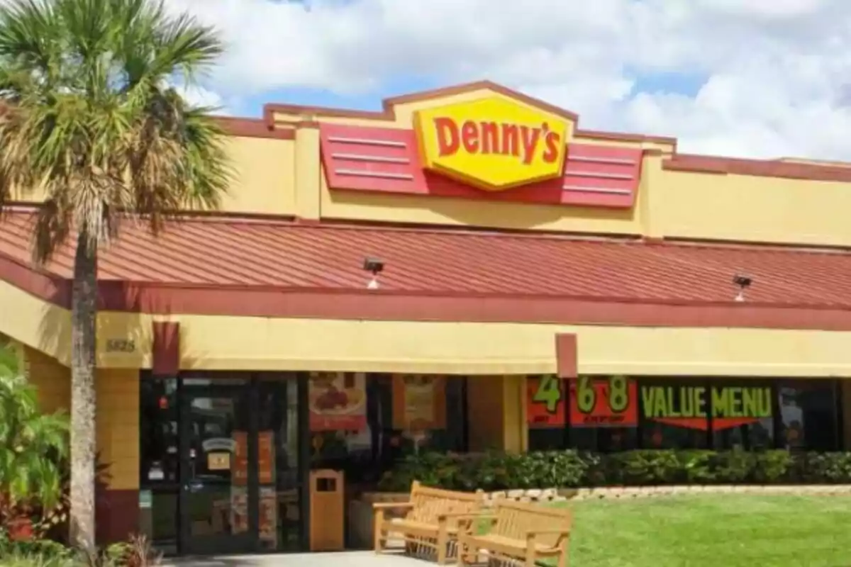 Fachada de un restaurante Denny's con un letrero amarillo y rojo y un área de asientos al aire libre rodeada de vegetación. Fachada de un restaurante Denny's con un letrero amarillo y rojo y un área de asientos al aire libre rodeada de vegetación.