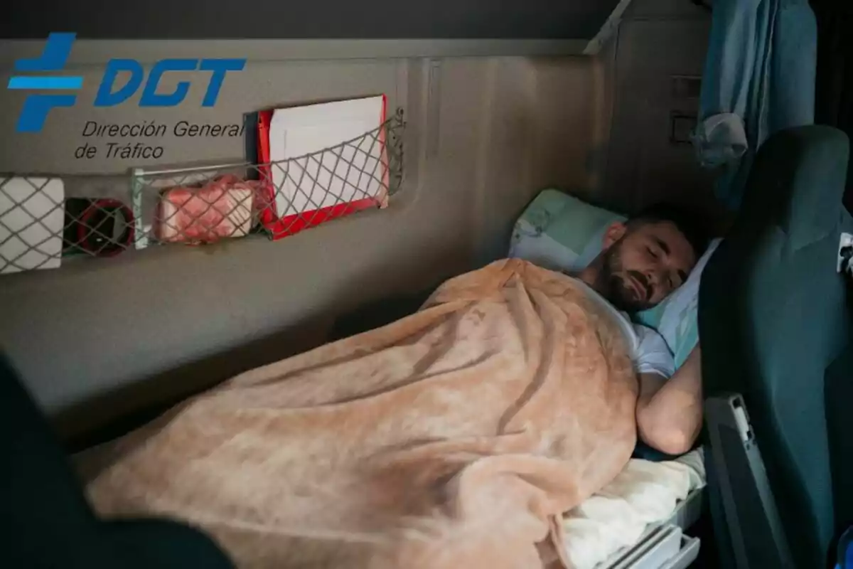 Un home dormint a la cabina d'un camió amb una manta beix, amb el logotip de la Direcció General de Trànsit (DGT) a la cantonada superior esquerra.