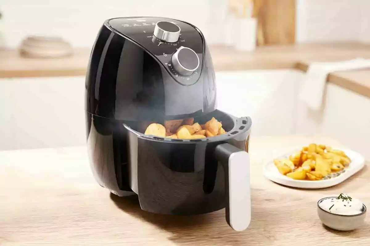 Fregidora d’aire negra cuinant patates en una cuina moderna amb plats de patates i salsa al fons Fregidora d’aire negra cuinant patates en una cuina moderna amb plats de patates i salsa al fons