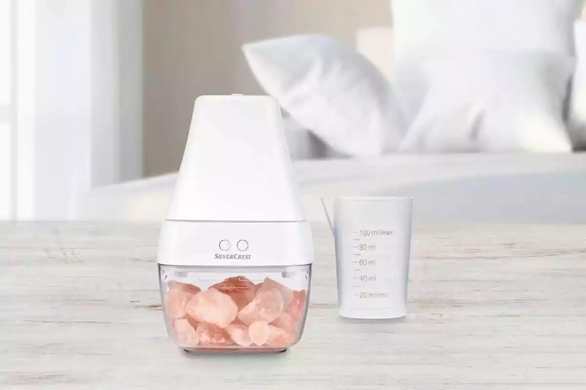 Un difusor blanc de Lidl amb pedres rosades a la base al costat d'un got mesurador transparent sobre una superfície de fusta clara en un entorn de dormitori. Un difusor blanc de Lidl amb pedres rosades a la base al costat d'un got mesurador transparent sobre una superfície de fusta clara en un entorn de dormitori.