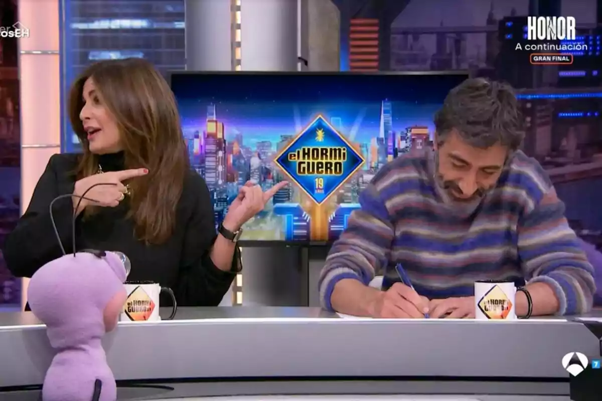 En un estudio de televisión, Nuria Roca gesticula mientras Juan del Val escribe, con un muñeco morado en primer plano y un logotipo de 