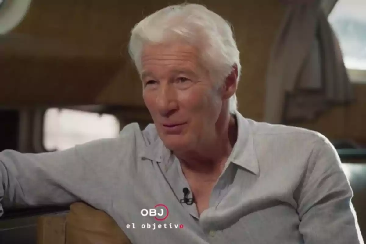 Richard Gere in a light jersey is sitting and speaking indoors with curtains in the background. Screenshot from 'El Objetivo'. Richard Gere in a light jersey is sitting and speaking indoors with curtains in the background. Screenshot from 'El Objetivo'.