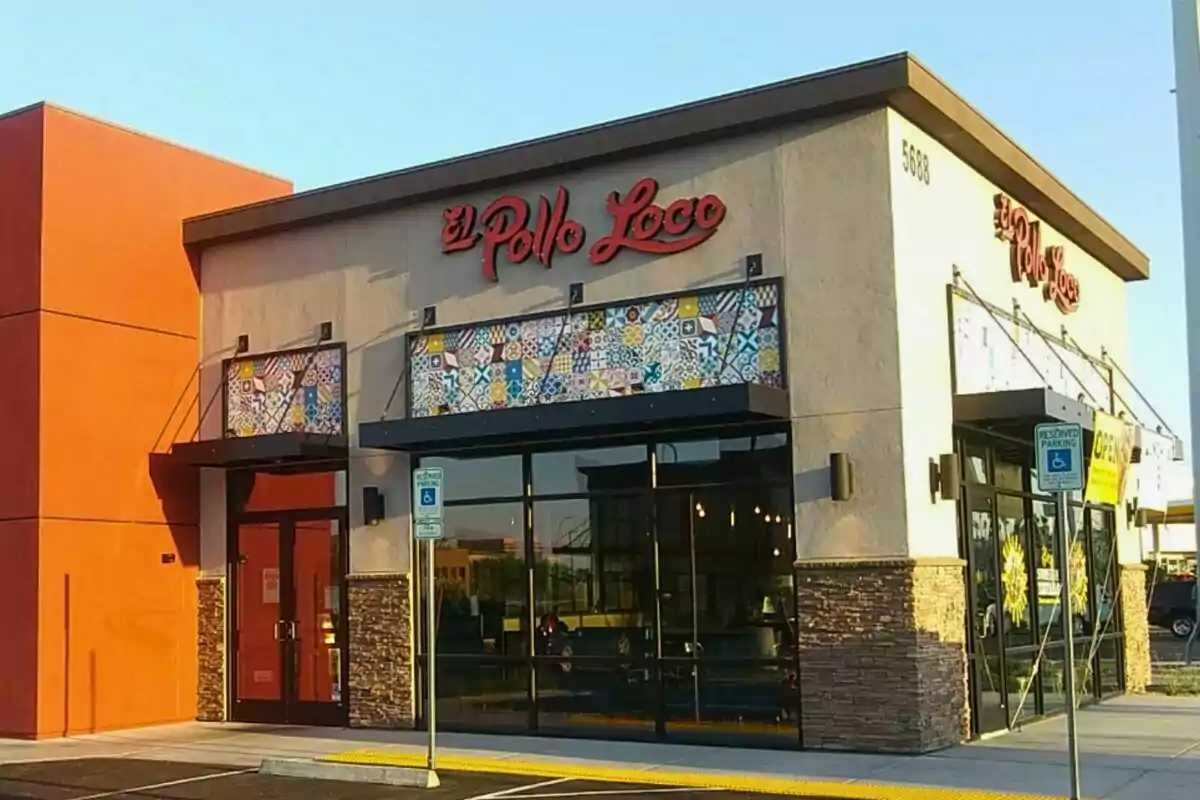 Façana d’un restaurant El Pollo Loco amb parets beix i detalls taronja, finestres grans i rètol vermell a la part superior Façana d’un restaurant El Pollo Loco amb parets beix i detalls taronja, finestres grans i rètol vermell a la part superior