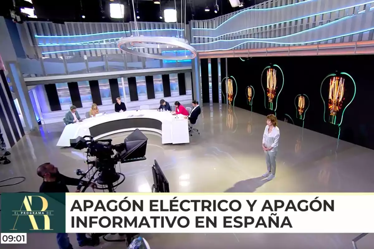 Un estudi de televisió amb diverses persones assegudes en una taula corba i una persona dreta, amb càmeres i llums al sostre, i un fons de bombetes il·luminades. Captura de 'El Programa de AR' Un estudi de televisió amb diverses persones assegudes en una taula corba i una persona dreta, amb càmeres i llums al sostre, i un fons de bombetes il·luminades. Captura de 'El Programa de AR'