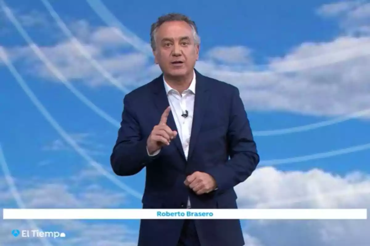 Un home amb vestit fosc està dret davant d'un fons blau amb núvols i línies blanques, gesticulant amb una mà mentre parla. Un home amb vestit fosc està dret davant d'un fons blau amb núvols i línies blanques, gesticulant amb una mà mentre parla.