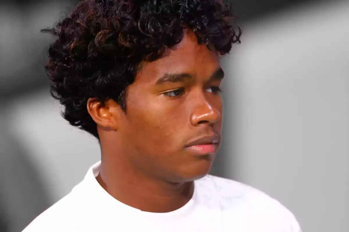 Young man with curly hair and serious expression, wearing a white t-shirt.