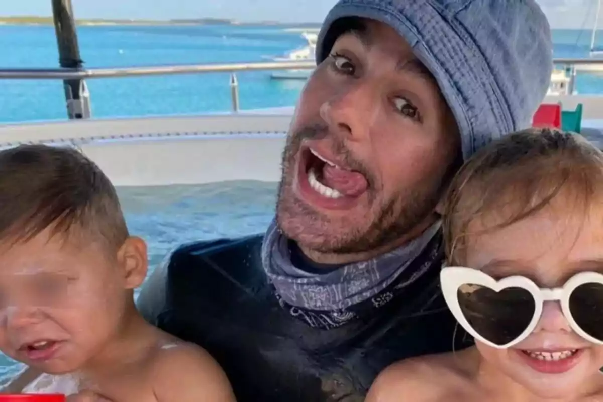 Una persona con gorro y bufanda está haciendo una mueca divertida mientras sostiene a dos niños pequeños, uno de los cuales lleva gafas de sol en forma de corazón, en un entorno acuático con un paisaje marino de fondo. Una persona con gorro y bufanda está haciendo una mueca divertida mientras sostiene a dos niños pequeños, uno de los cuales lleva gafas de sol en forma de corazón, en un entorno acuático con un paisaje marino de fondo.