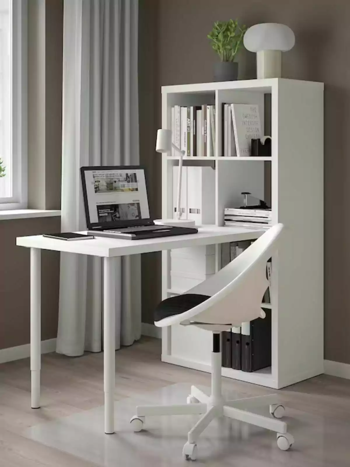 Espacio de trabajo moderno con escritorio blanco de Ikea con una silla giratoria y una estantería con libros y laptop frente a una ventana con cortinas blancas.