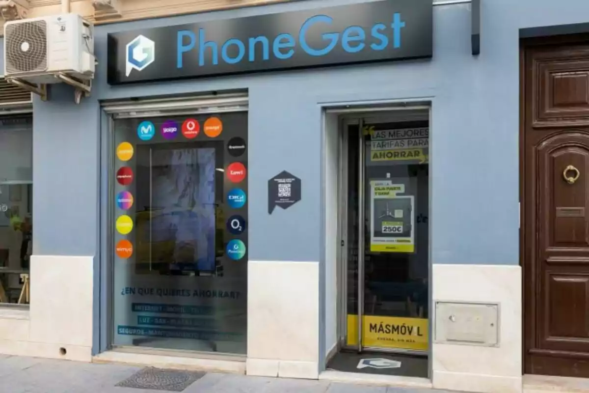 Façana d'una botiga de telefonia anomenada PhoneGest amb logotips de diverses companyies de telecomunicacions a la finestra i un cartell promocional a la porta.