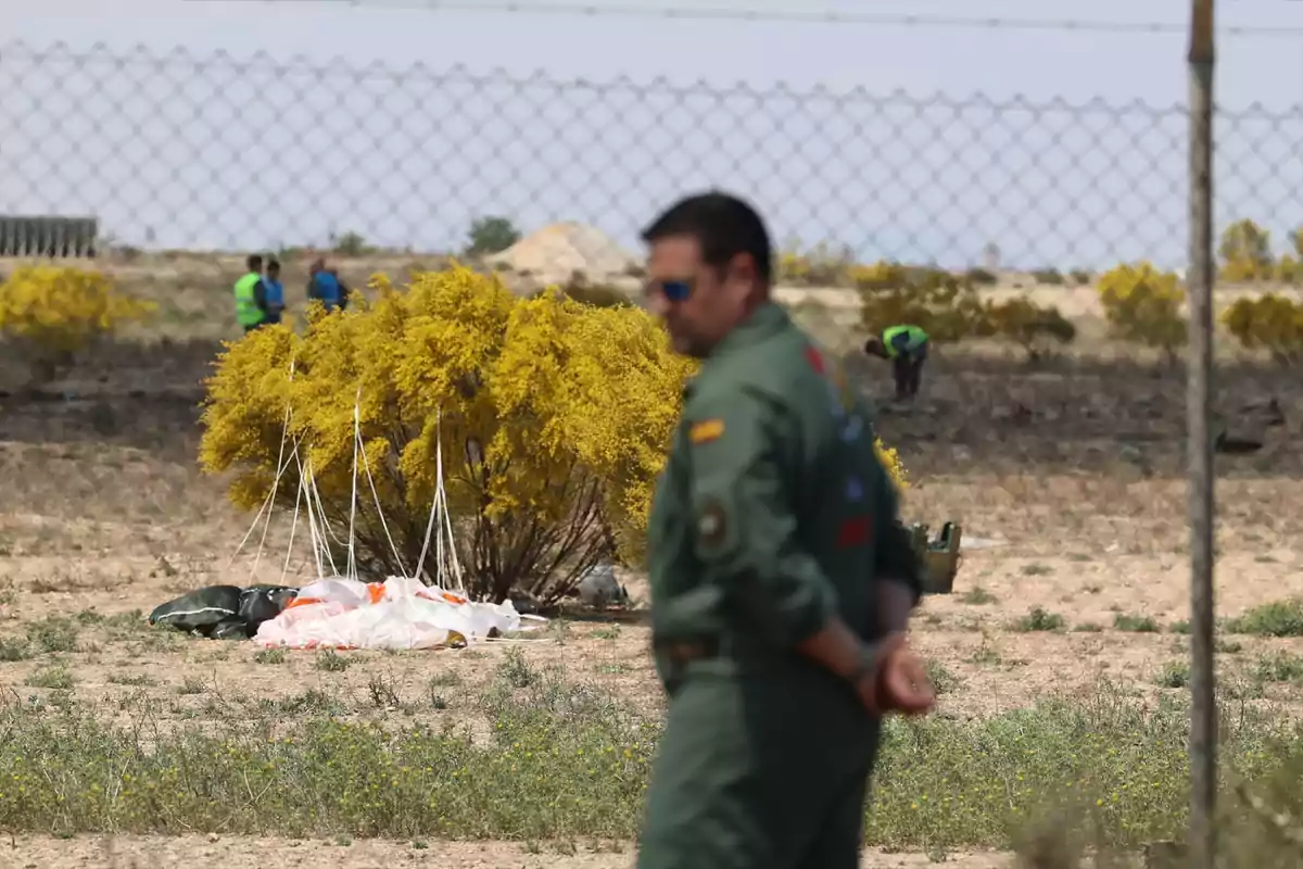 Un home amb uniforme militar està dret en un camp amb un paracaigudes a terra i arbustos grocs al fons.