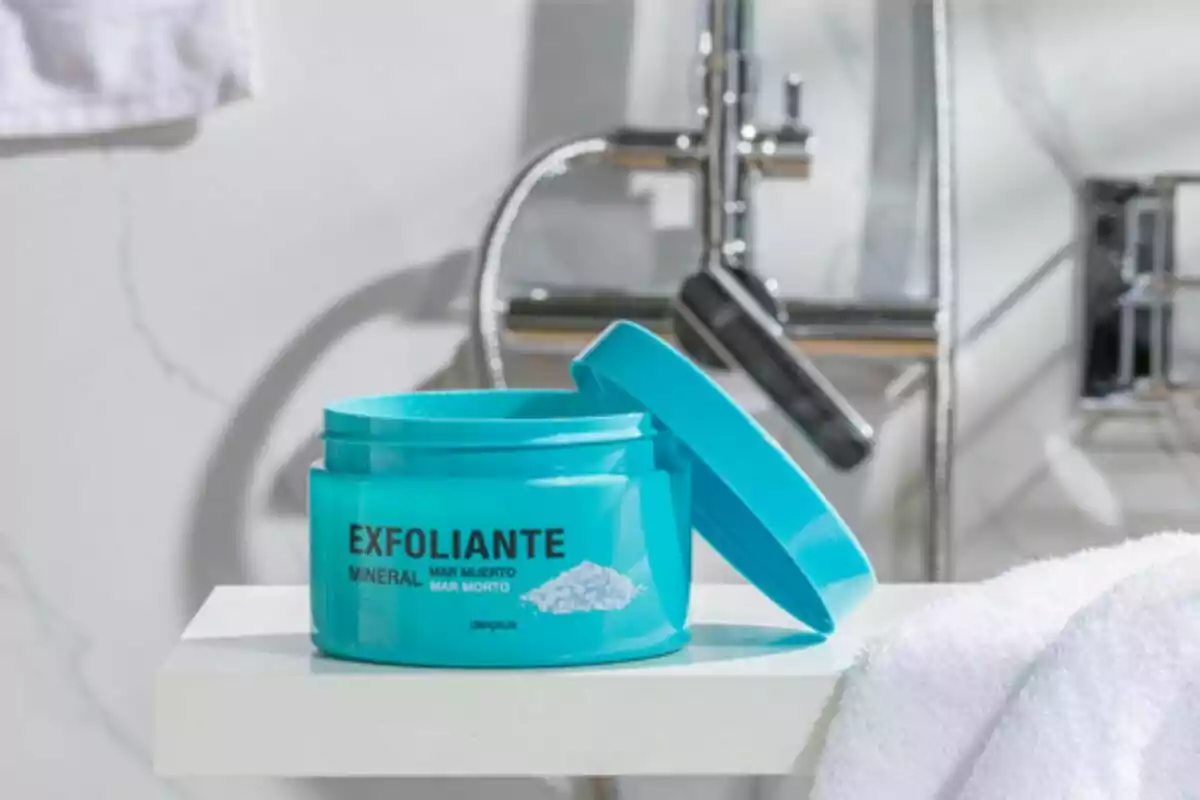 Envàs blau d'exfoliant mineral sobre una lleixa en un bany amb aixeta de fons.