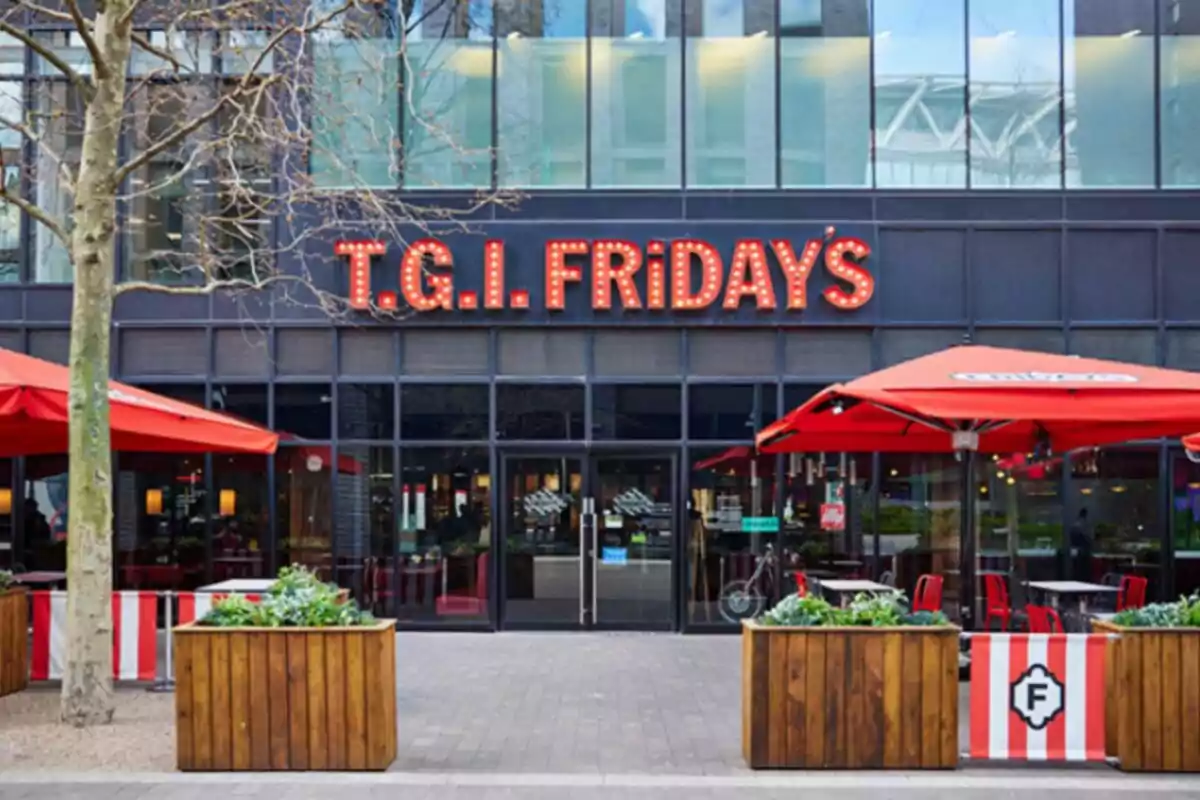 The image shows the facade of a T.G.I. Fridays restaurant with an illuminated sign and red umbrellas in the outdoor area. The image shows the facade of a T.G.I. Fridays restaurant with an illuminated sign and red umbrellas in the outdoor area.