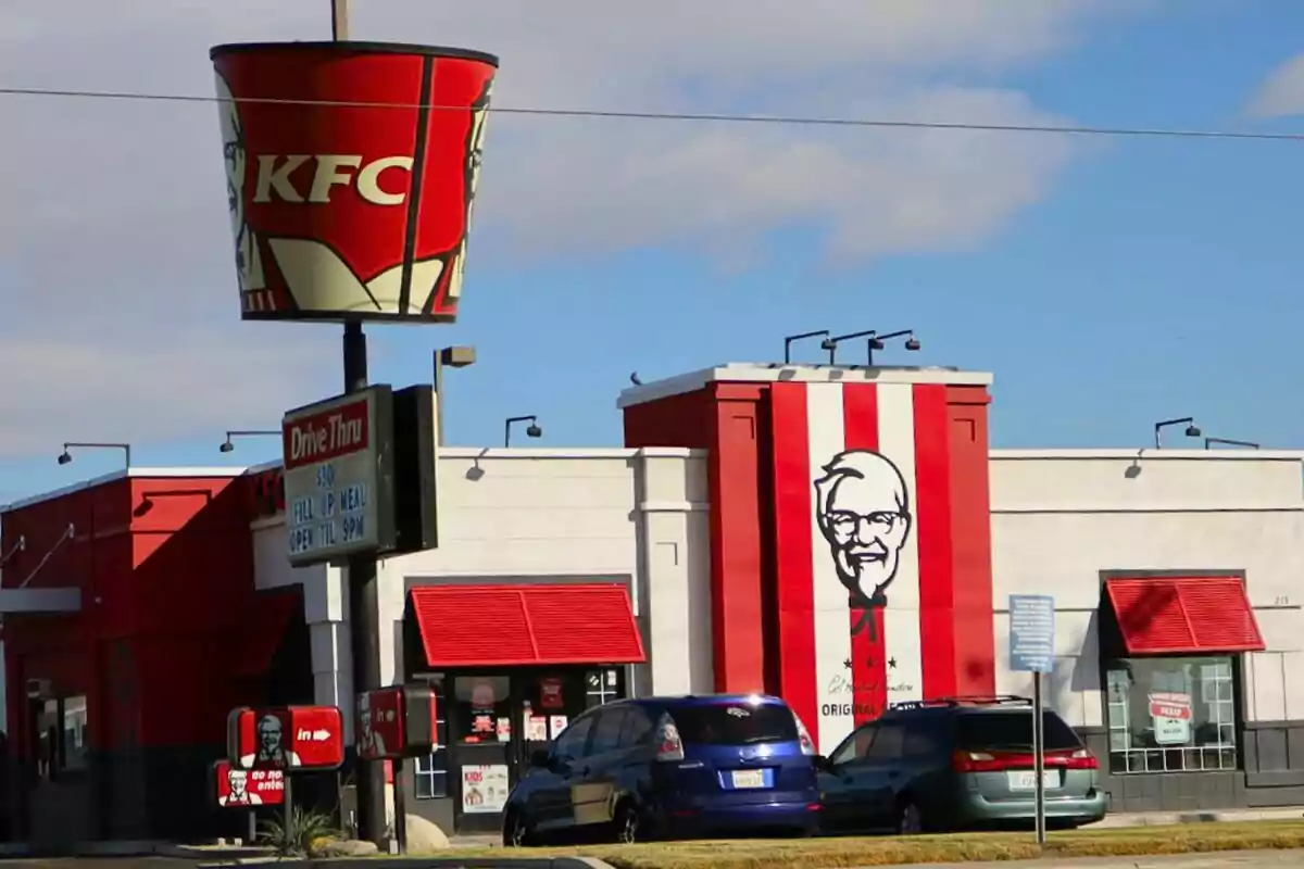 Un restaurant de menjar ràpid amb un gran cubell de pollastre fregit com a rètol -que és KFC- i un mural del fundador a la façana.