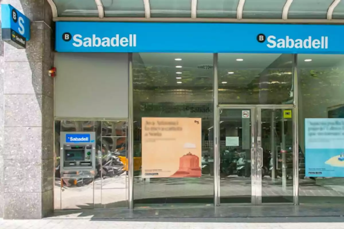 Façana d'una sucursal del banc Sabadell amb un caixer automàtic a l'esquerra i dos cartells publicitaris a les finestres.