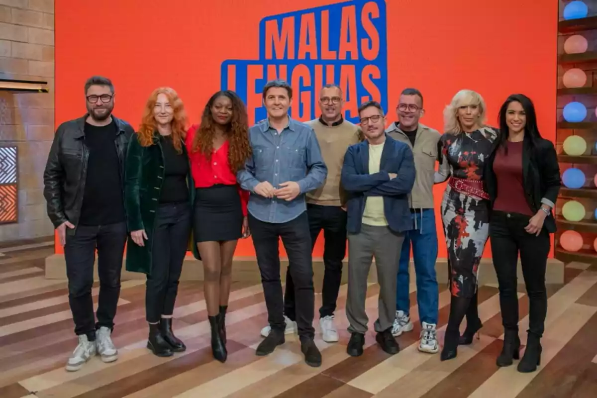 Un grupo de personas posando juntas en un set de televisión con un fondo naranja que tiene el texto Un grupo de personas posando juntas en un set de televisión con un fondo naranja que tiene el texto