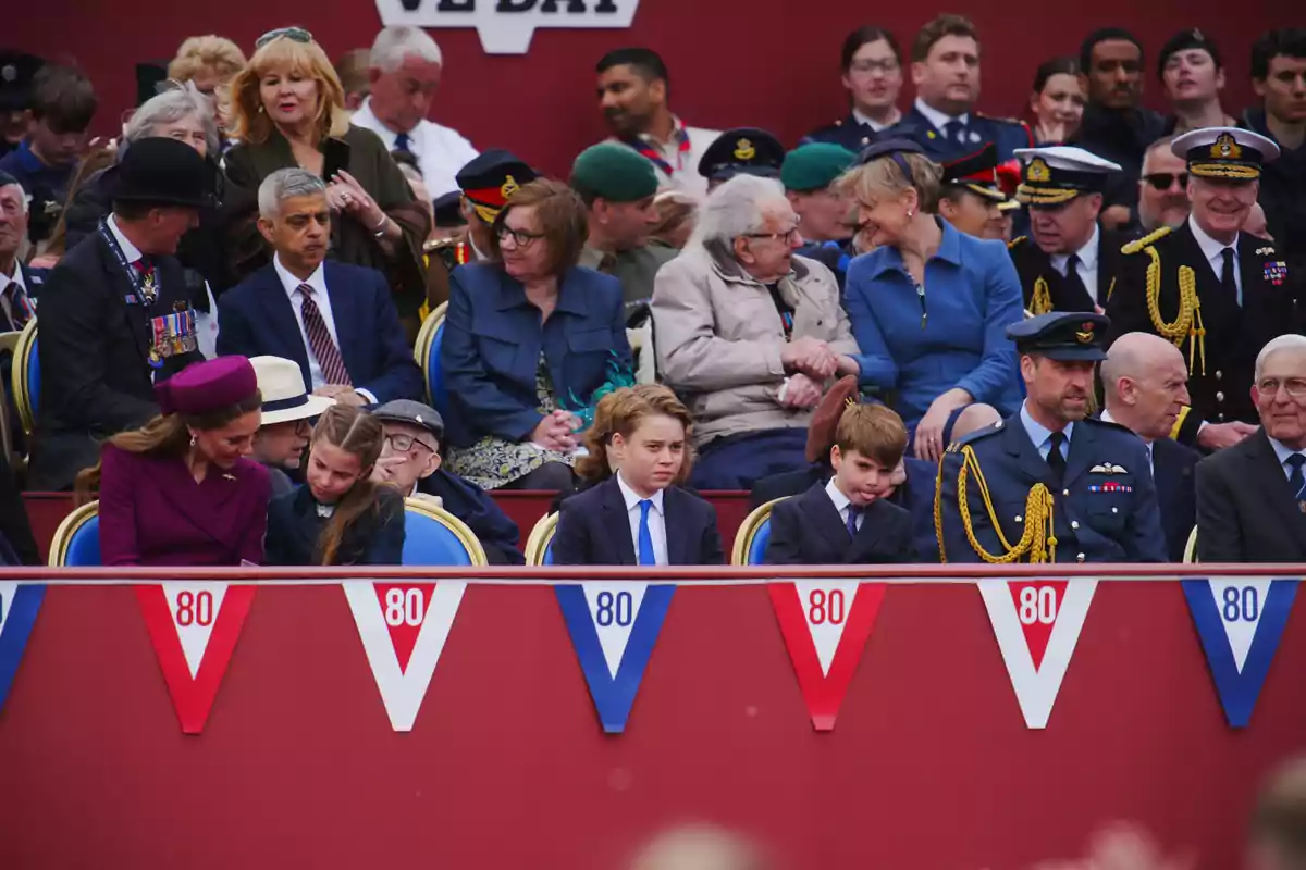 A group of people, some in military uniforms, are seated in a stand decorated with red, white, and blue pennants with the number 80, as they watch an event. A group of people, some in military uniforms, are seated in a stand decorated with red, white, and blue pennants with the number 80, as they watch an event.