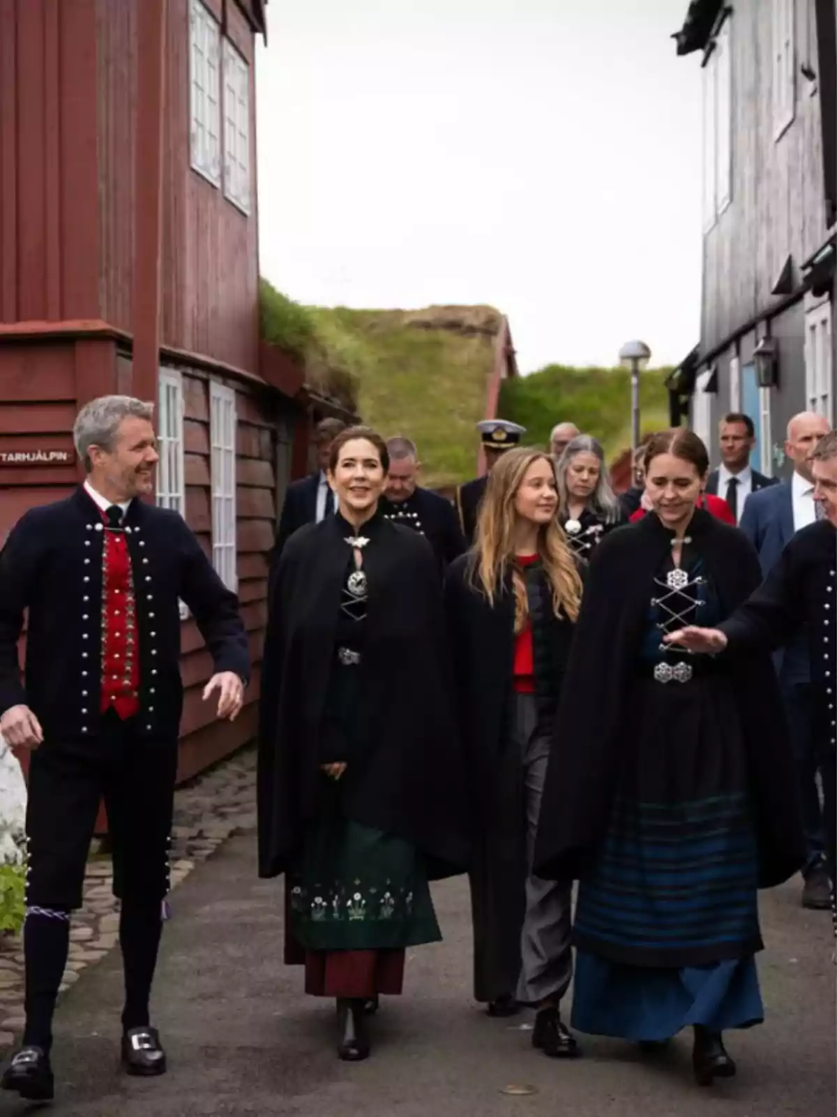 Grup de persones caminant per un carrer entre cases de fusta, algunes d’elles porten vestits tradicionals i capes fosques Grup de persones caminant per un carrer entre cases de fusta, algunes d’elles porten vestits tradicionals i capes fosques