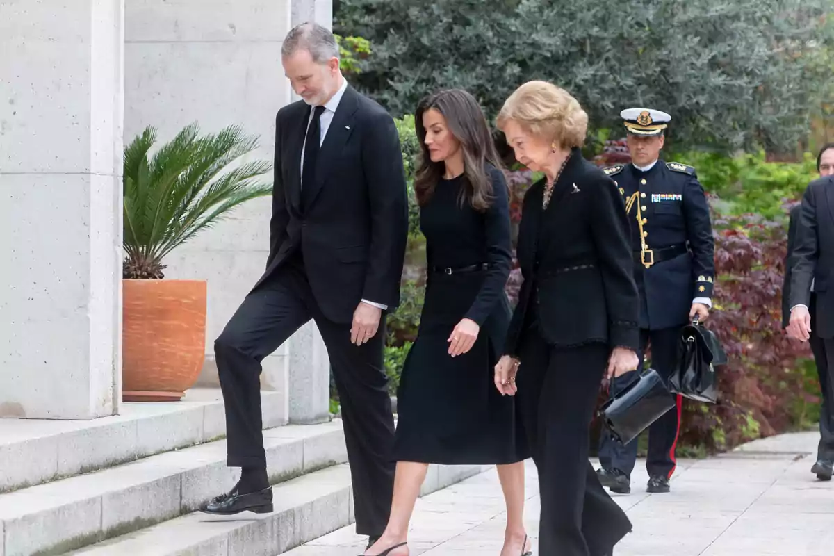 Felipe VI, Queen Letizia, and Queen Sofía, dressed in black, walk up some stairs outdoors accompanied by a man in military uniform. Felipe VI, Queen Letizia, and Queen Sofía, dressed in black, walk up some stairs outdoors accompanied by a man in military uniform.