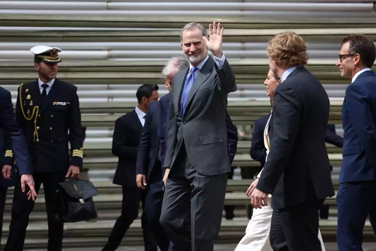 Felip VI amb vestit gris i corbata blava saluda amb la mà aixecada mentre camina acompanyat de diverses persones vestides formalment, incloent-hi un oficial naval. Felip VI amb vestit gris i corbata blava saluda amb la mà aixecada mentre camina acompanyat de diverses persones vestides formalment, incloent-hi un oficial naval.