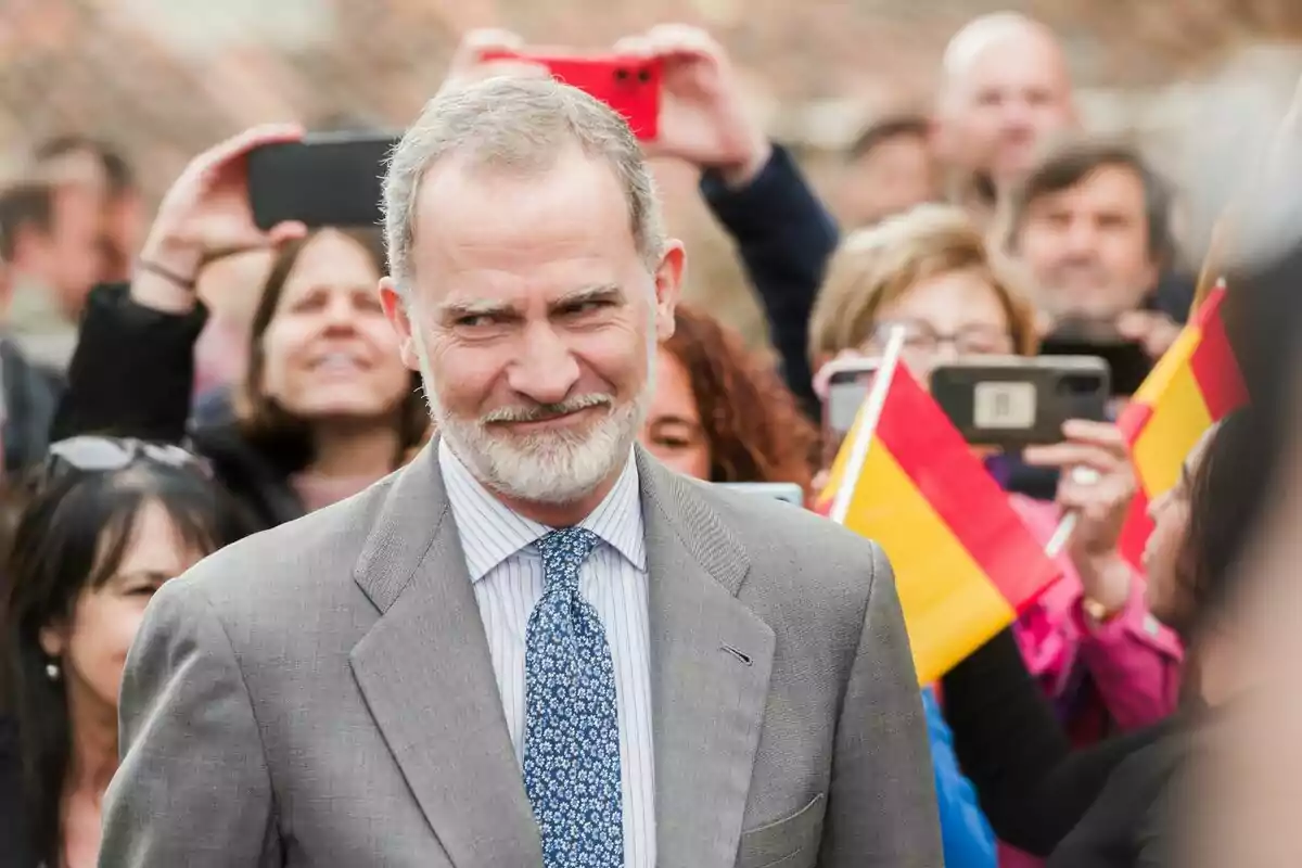 Felip VI amb vestit gris i corbata blava somriu mentre una multitud el fotografia i oneja banderes d’Espanya. Felip VI amb vestit gris i corbata blava somriu mentre una multitud el fotografia i oneja banderes d’Espanya.