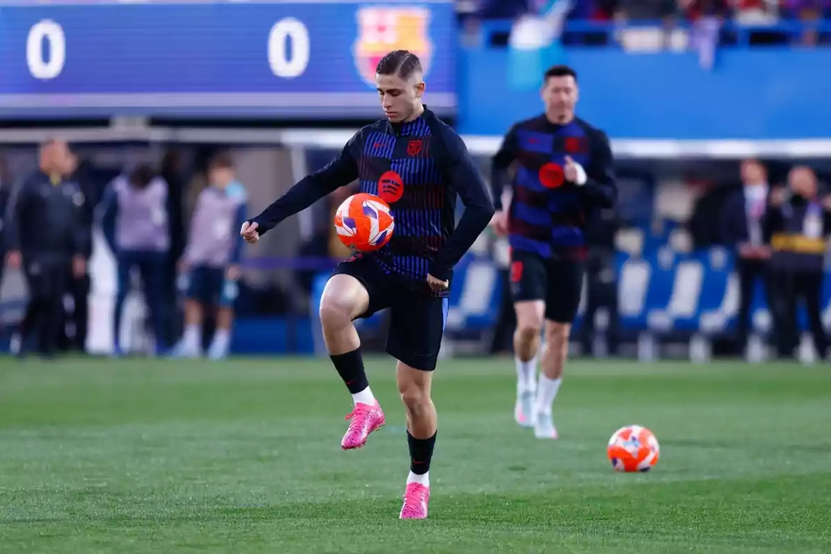 A player in a dark uniform and pink cleats controls a ball on the field while another player runs behind him.