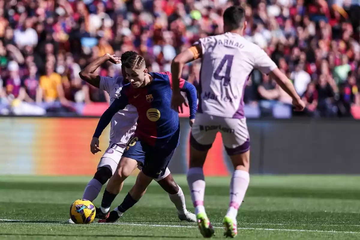 A player in a blue and red uniform controls the ball while being marked by opponents in white and purple uniforms in a stadium full of spectators.