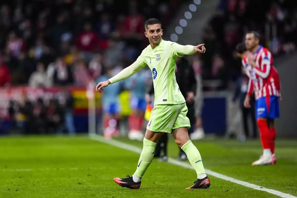 Un jugador de futbol amb uniforme verd clar somriu i assenyala cap a un costat en un camp de joc, mentre un altre jugador amb uniforme vermell i blanc es troba al fons. Un jugador de futbol amb uniforme verd clar somriu i assenyala cap a un costat en un camp de joc, mentre un altre jugador amb uniforme vermell i blanc es troba al fons.