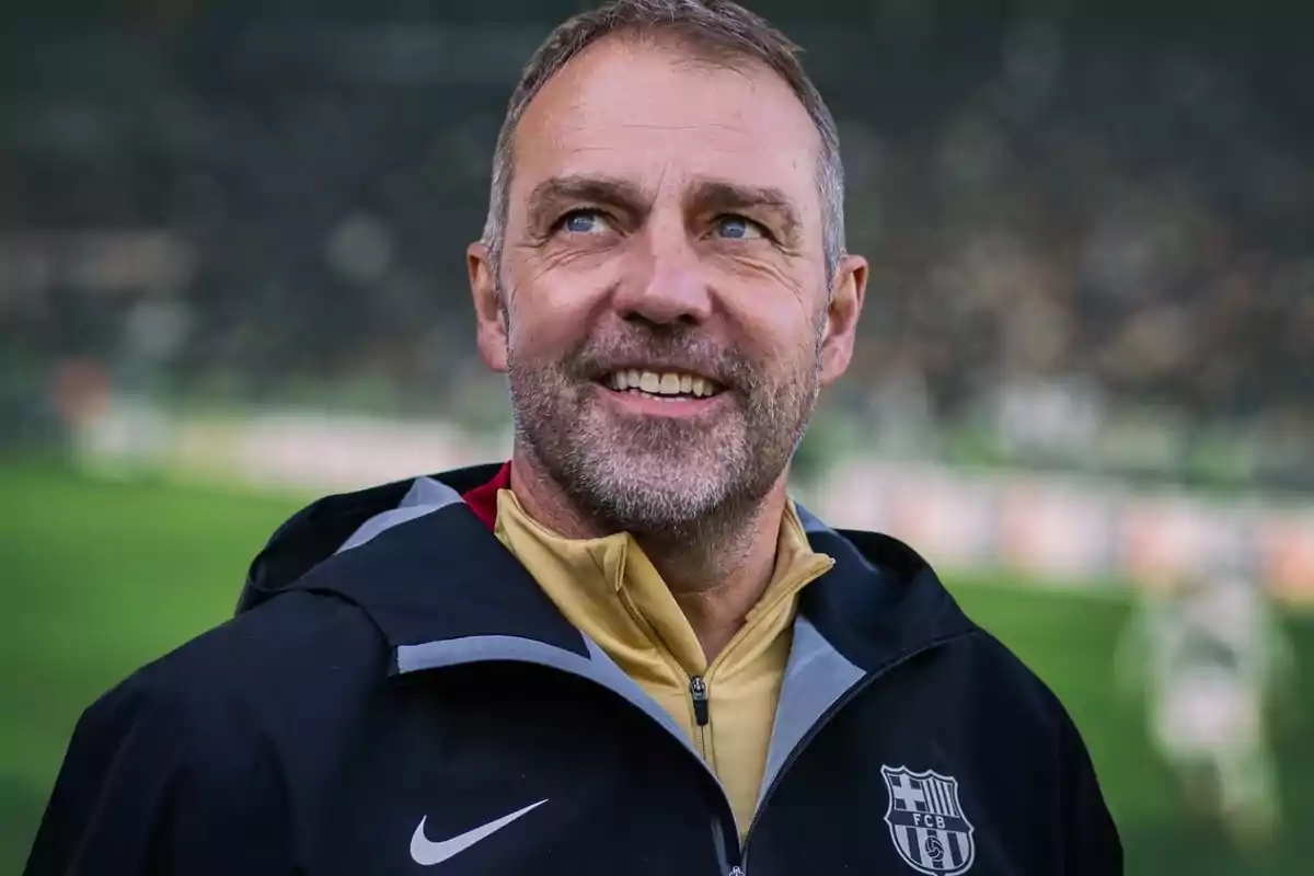 A smiling man with a beard and short hair wears a sports jacket with a soccer team logo on a blurred green field. A smiling man with a beard and short hair wears a sports jacket with a soccer team logo on a blurred green field.
