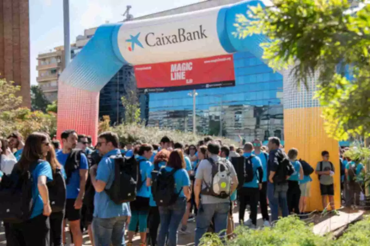Un grup de persones amb samarretes blaves i motxilles es reuneix sota un arc inflable de CaixaBank en un esdeveniment a l'aire lliure. Un grup de persones amb samarretes blaves i motxilles es reuneix sota un arc inflable de CaixaBank en un esdeveniment a l'aire lliure.