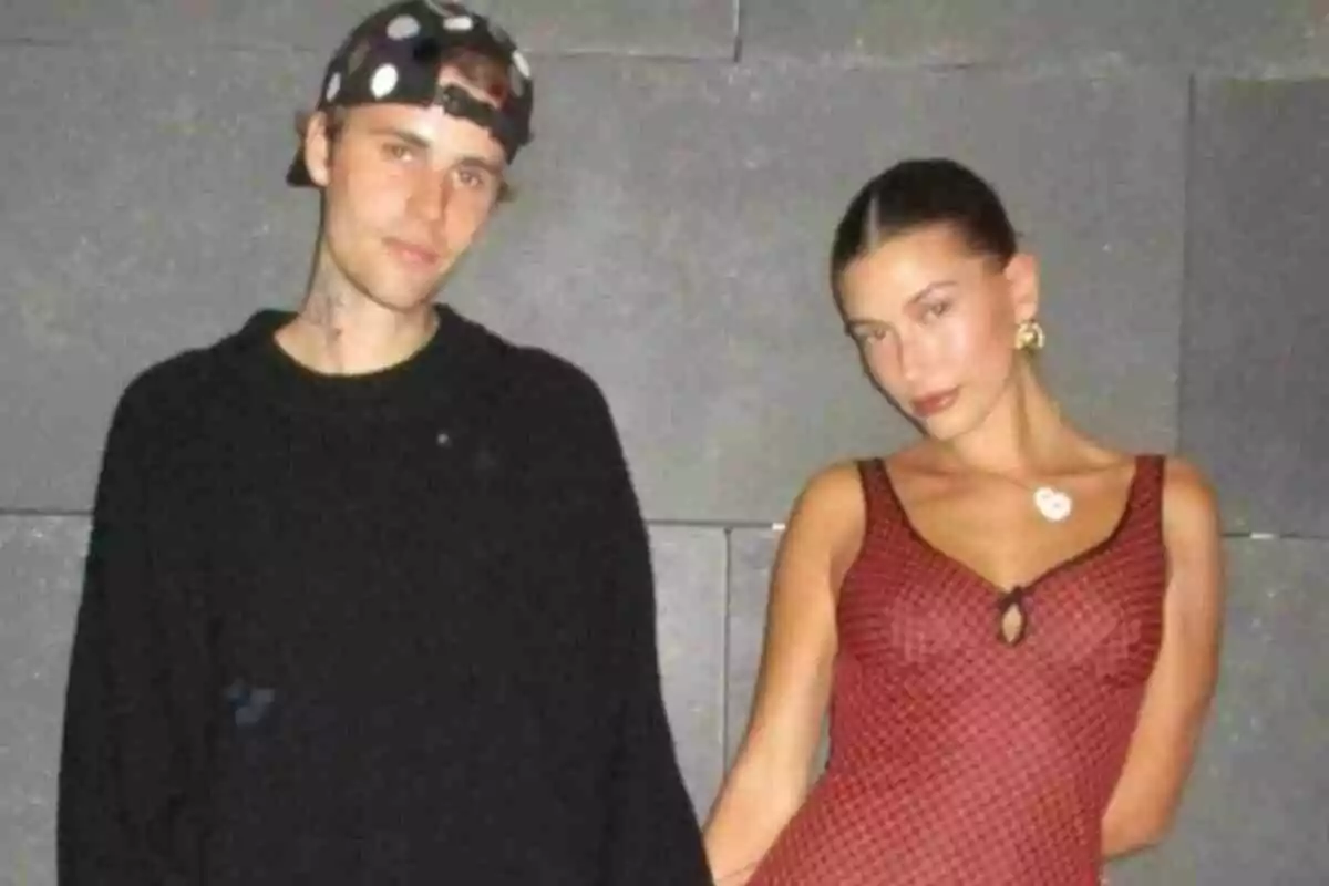Justin Bieber and Hailey Bieber posing together in front of a gray wall, he's wearing a black sweater and cap, she's a red dress with a pattern. Justin Bieber and Hailey Bieber posing together in front of a gray wall, he's wearing a black sweater and cap, she's a red dress with a pattern.