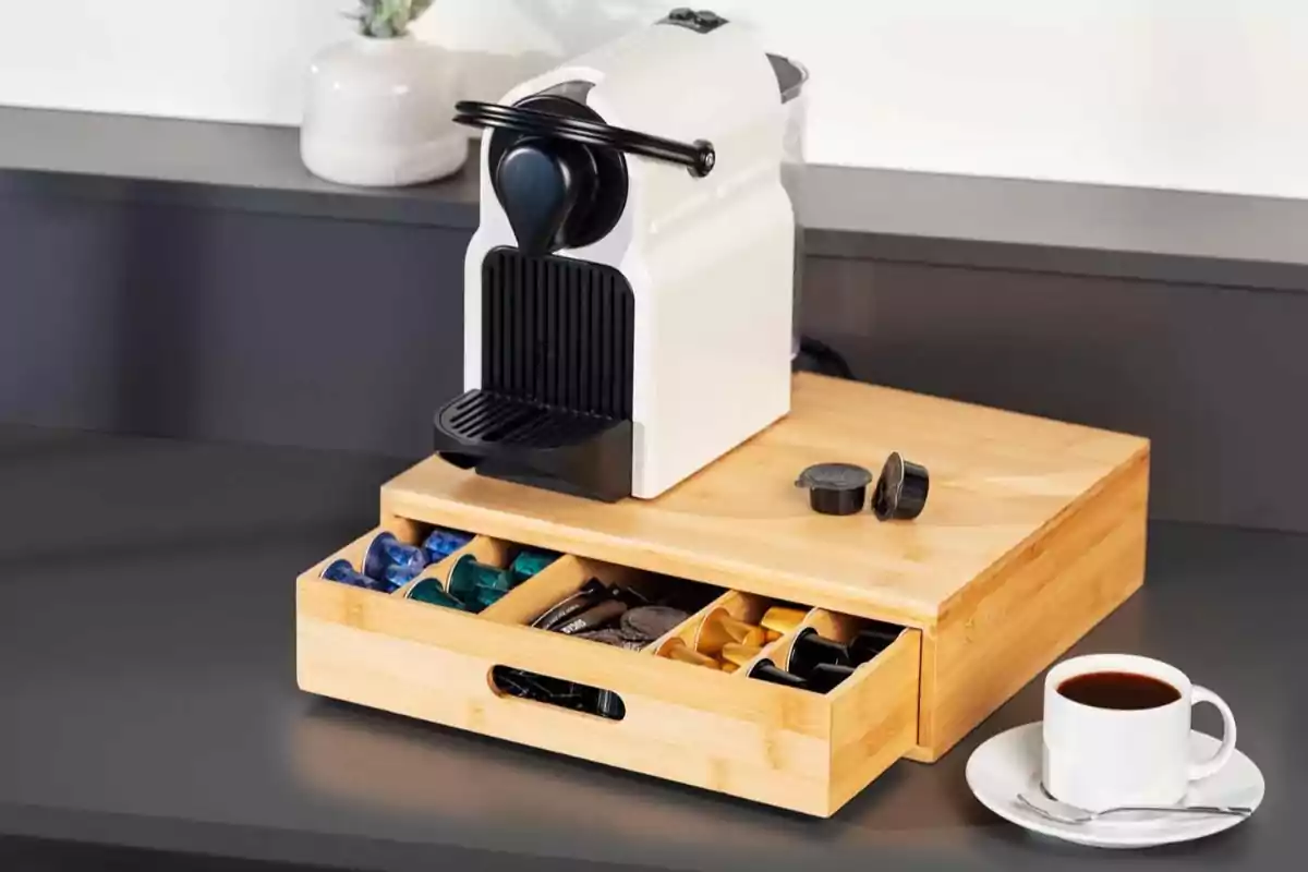 Cafetera de càpsules blanca sobre una caixa organitzadora de fusta amb càpsules de cafè i una tassa de cafè sobre una taula Cafetera de càpsules blanca sobre una caixa organitzadora de fusta amb càpsules de cafè i una tassa de cafè sobre una taula