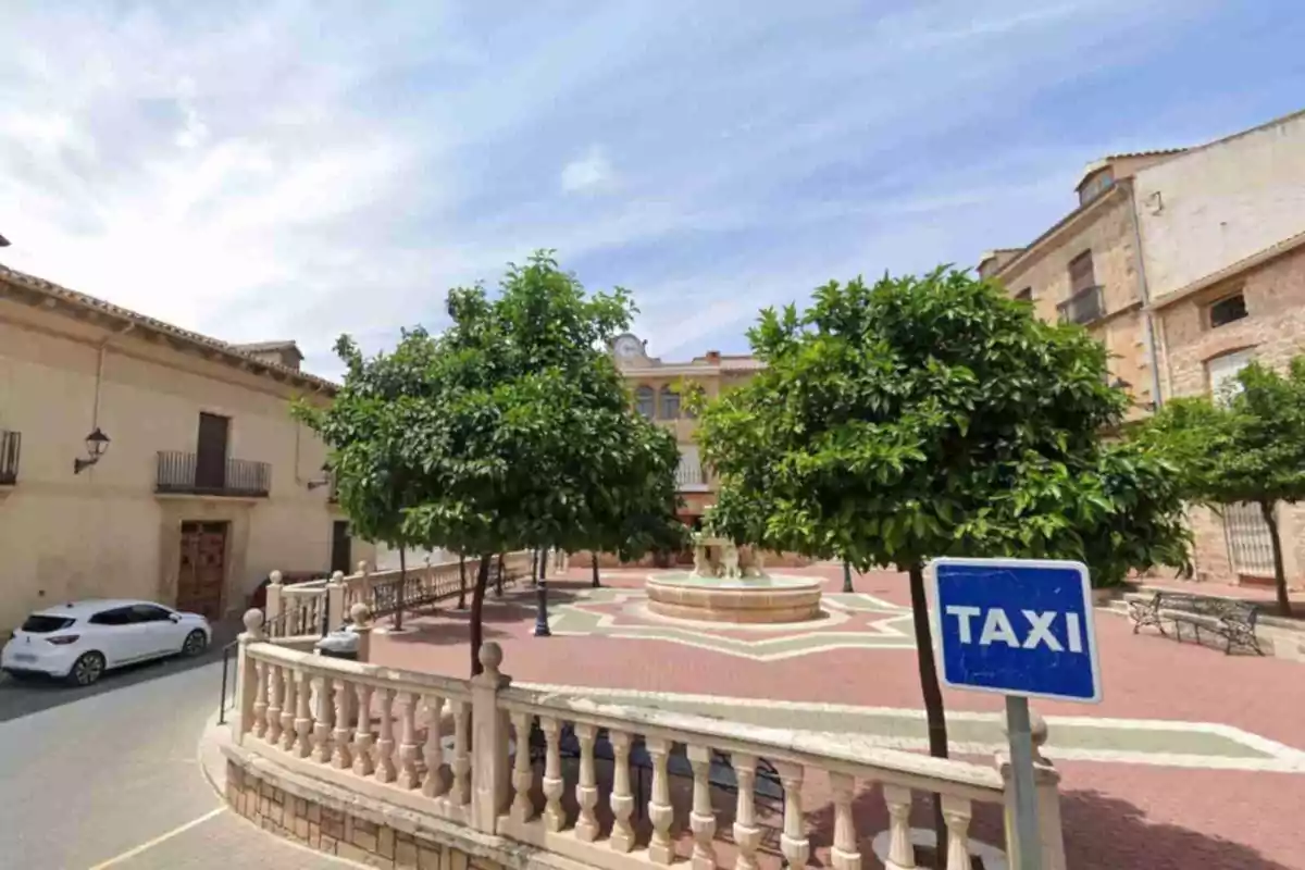 Plaça amb arbres, una font central, bancs i un rètol blau de taxi en primer pla a Sorihuela del Guadalimar Plaça amb arbres, una font central, bancs i un rètol blau de taxi en primer pla a Sorihuela del Guadalimar