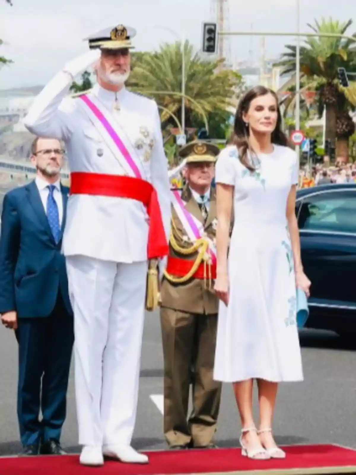 Un home amb uniforme militar blanc saluda mentre una dona amb vestit blanc està dreta al seu costat sobre una catifa vermella en un acte oficial a l’aire lliure. Un home amb uniforme militar blanc saluda mentre una dona amb vestit blanc està dreta al seu costat sobre una catifa vermella en un acte oficial a l’aire lliure.