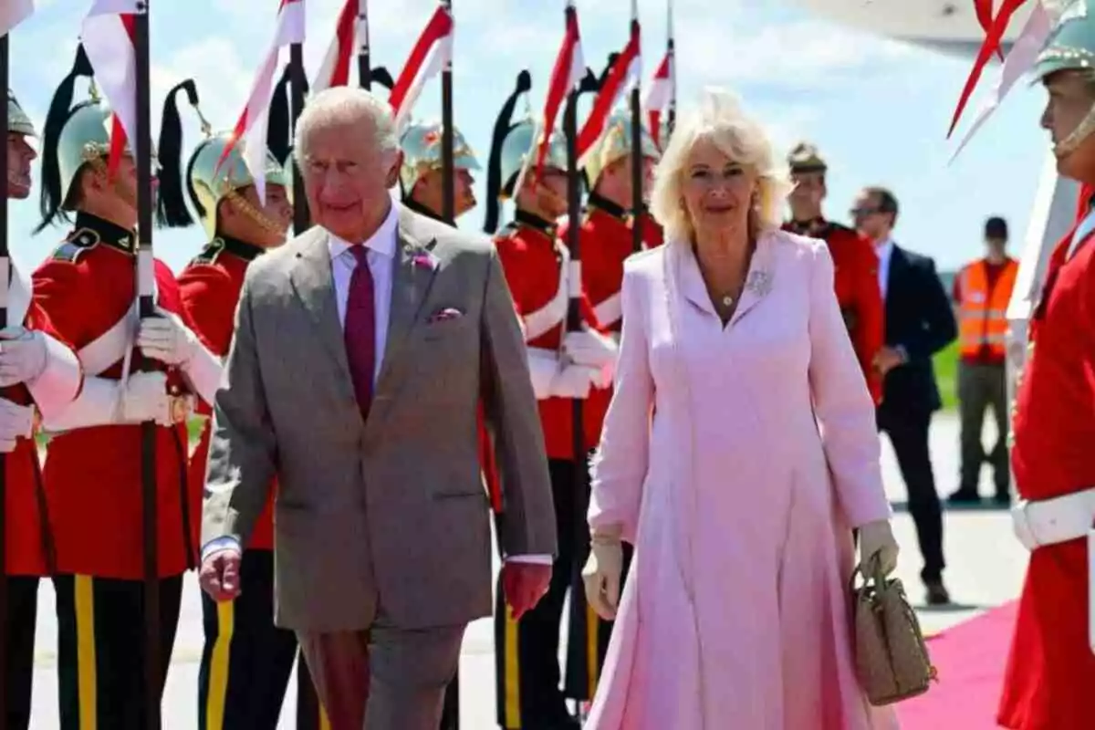 King Charles III and Queen Camila elegantly dressed walk down a red carpet while soldiers in red uniforms and silver helmets form a guard of honor around them. King Charles III and Queen Camila elegantly dressed walk down a red carpet while soldiers in red uniforms and silver helmets form a guard of honor around them.