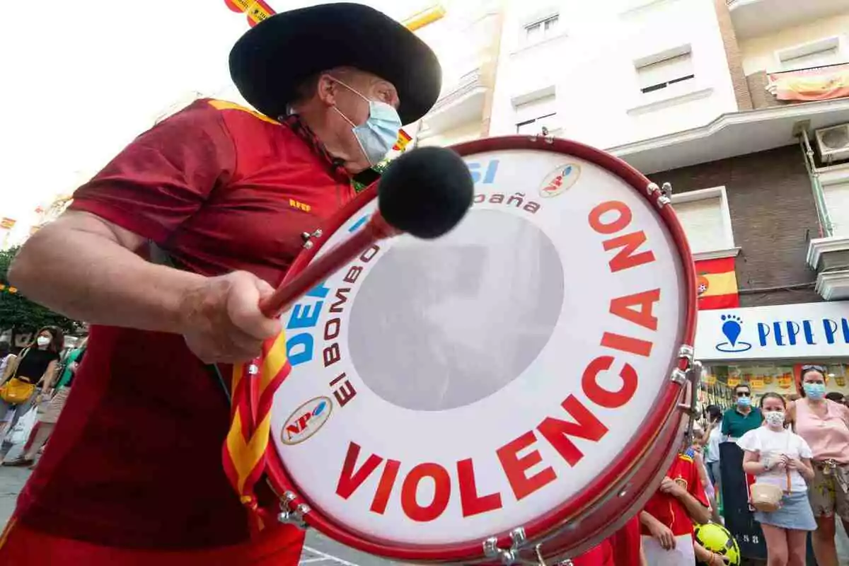 Manolo El del Bombo amb barret i mascareta toca un bombo que té un missatge contra la violència mentre persones observen al carrer. Manolo El del Bombo amb barret i mascareta toca un bombo que té un missatge contra la violència mentre persones observen al carrer.