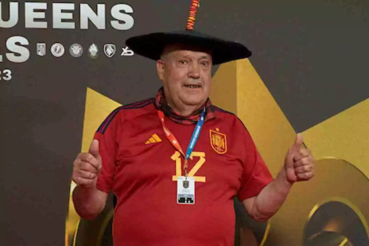 Manolo El del Bombo amb samarreta vermella d'Espanya i barret negre somriu i aixeca els polzes davant d'un fons decoratiu. Manolo El del Bombo amb samarreta vermella d'Espanya i barret negre somriu i aixeca els polzes davant d'un fons decoratiu.