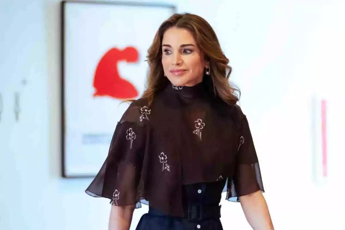 Rania of Jordan with wavy brown hair is wearing a black blouse with floral details and is standing in front of a white wall with a painting in the background. Rania of Jordan with wavy brown hair is wearing a black blouse with floral details and is standing in front of a white wall with a painting in the background.