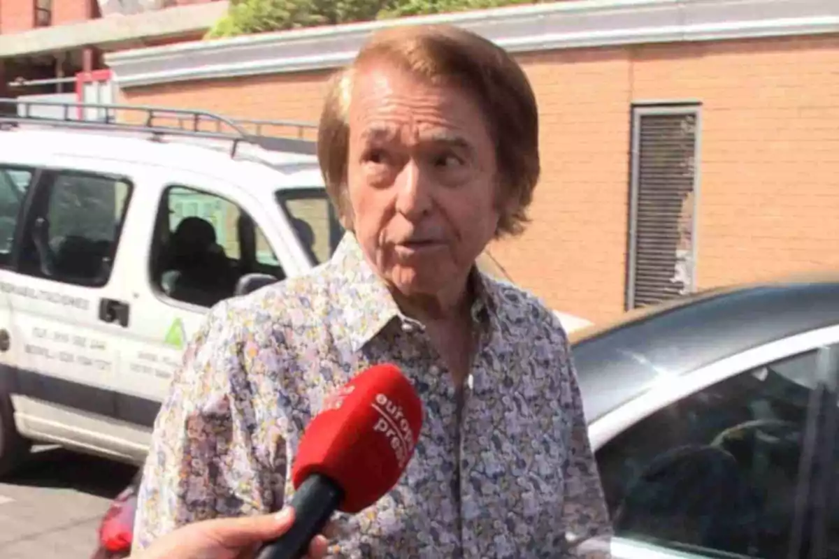 Raphael amb camisa estampada sent entrevistat al carrer davant d’una furgoneta blanca i un cotxe negre Raphael amb camisa estampada sent entrevistat al carrer davant d’una furgoneta blanca i un cotxe negre