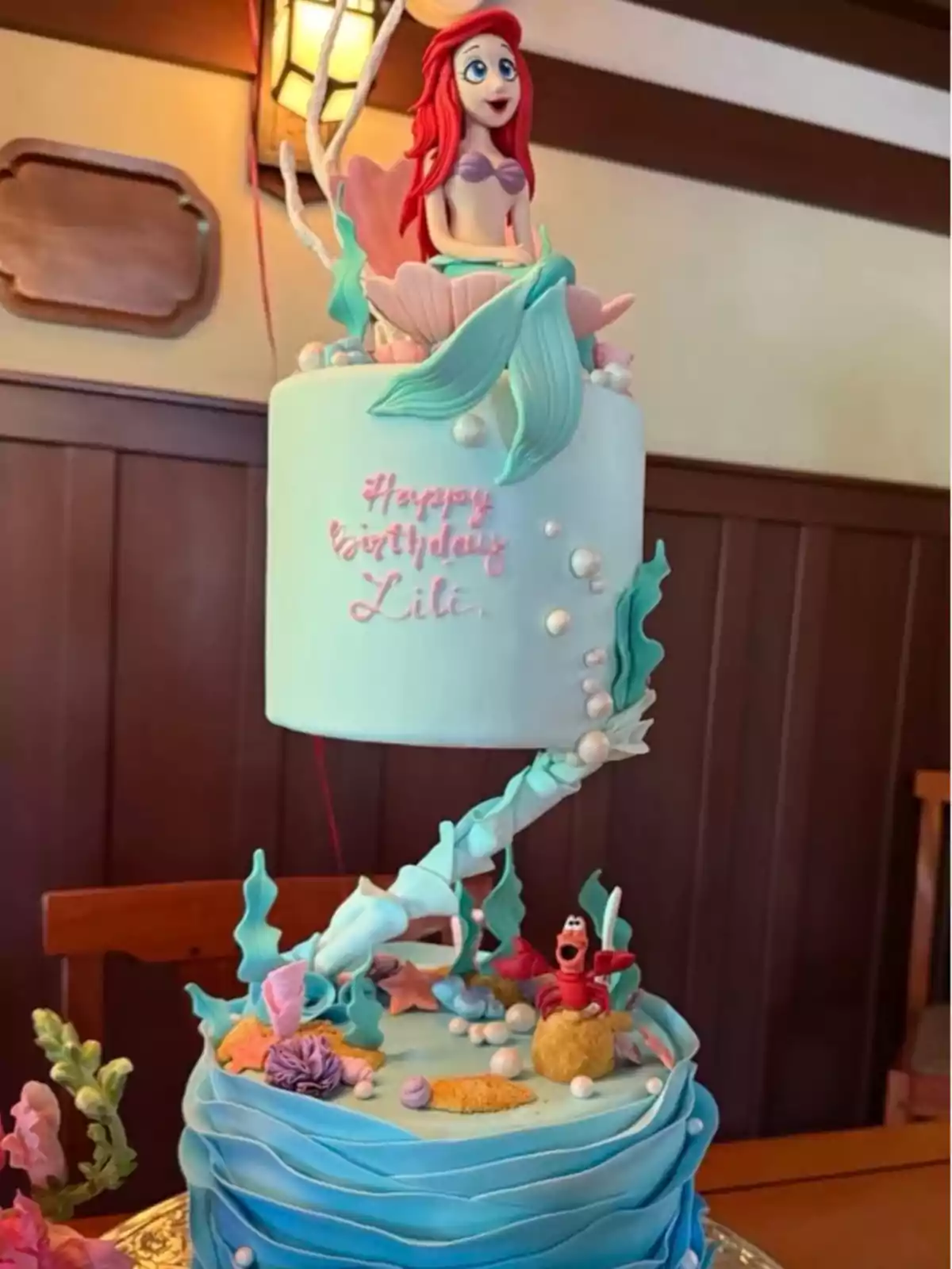 Pastel de dos pisos decorado con temática de La Sirenita, con una figura de Ariel en la parte superior y detalles marinos como corales, perlas y un cangrejo rojo en la base, además de un mensaje de cumpleaños para Lili