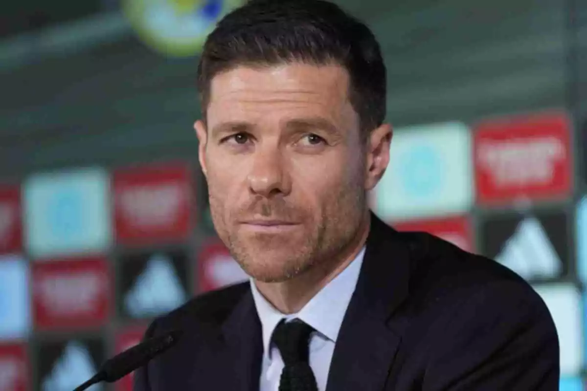 Xabi Alonso amb cabell curt i barba retallada amb vestit fosc en una roda de premsa amb un fons de logotips desenfocats Xabi Alonso amb cabell curt i barba retallada amb vestit fosc en una roda de premsa amb un fons de logotips desenfocats
