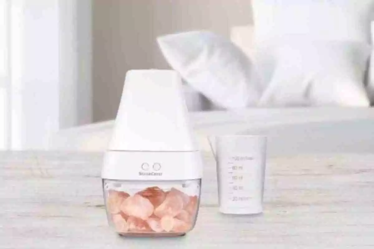 Un petit electrodomèstic blanc amb pedres rosades a l'interior al costat d'un got mesurador sobre una taula clara amb un llit desenfocat al fons. Un petit electrodomèstic blanc amb pedres rosades a l'interior al costat d'un got mesurador sobre una taula clara amb un llit desenfocat al fons.