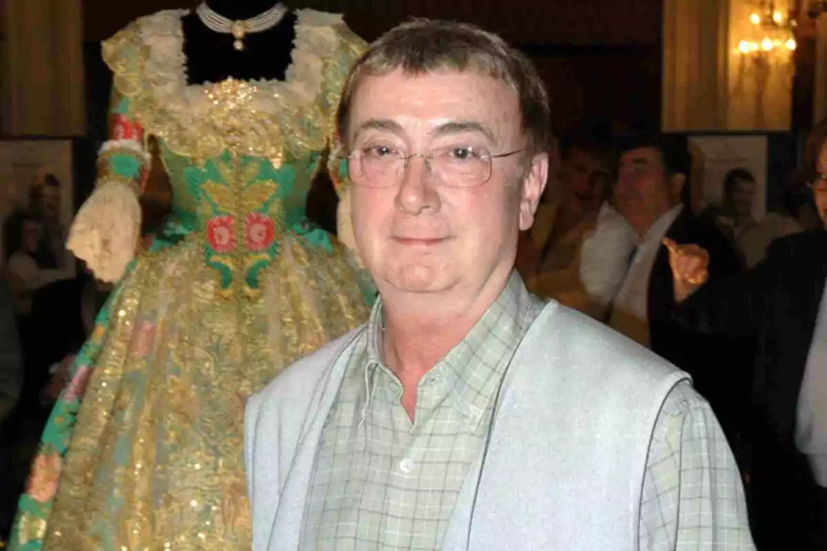 Enrique Marzal con cabello corto y gafas posando frente a un vestido tradicional de fallera colorido en un evento social.