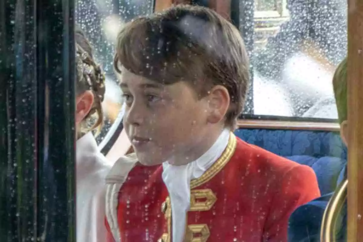 Prince George in a red uniform with gold details sits inside a vehicle, seen through a window with raindrops, in the coronation of King Charles III. Prince George in a red uniform with gold details sits inside a vehicle, seen through a window with raindrops, in the coronation of King Charles III.